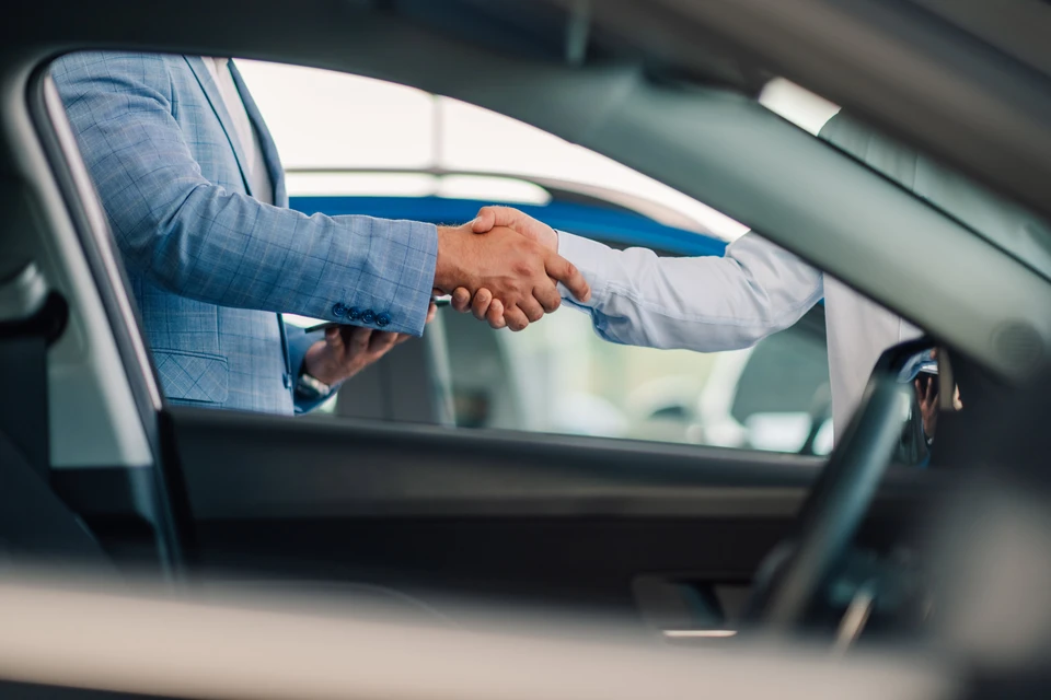Une poign&eacute;e de main finale et l'&eacute;change de cl&eacute;s de voiture entre deux individus, l'un portant un costume bleu et l'autre en chemise blanche, symbolisant la conclusion r&eacute;ussie d'une transaction chez un concessionnaire automobile