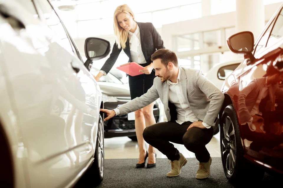 Concession automobile avec vendeur homme en costume examinant une voiture blanche, accompagné d'une vendeuse femme tenant des documents