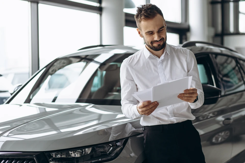 Un coach commercial en costume ou tenue professionnelle dynamique interagissant avec un vendeur automobile en concession automobile. Scène photographiée dans un showroom automobile moderne, avec des véhicules visibles en arrière-plan. Le coach et le vendeur sont en discussion engagée, le coach faisant des gestes pédagogiques, les deux personnages concentrés et professionnels. L'ambiance doit refléter un accompagnement technique et actif, pas une simple interaction sociale.