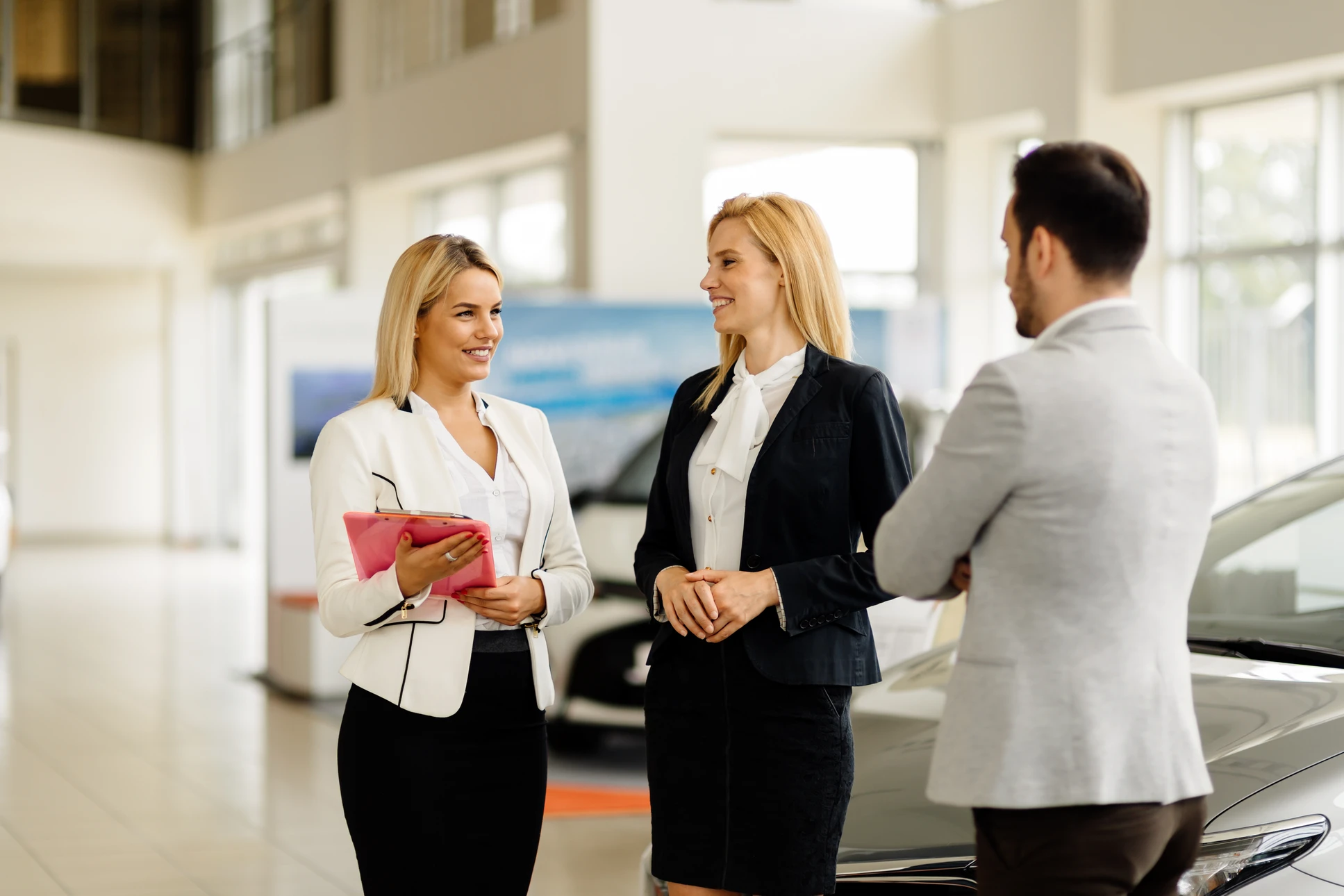 Photograph of a business coach or sales trainer leading a training session or coaching meeting with a team of car dealership sales professionals in a modern conference room or at a dealership showroom. The scene shows an experienced presenter engaging attentively with the group, with participants listening and taking notes. The setting reflects a professional automotive commercial environment with elements suggesting car sales (modern office, professional attire, sales-focused atmosphere). The image captures dynamic, constructive interaction between trainer and team members.