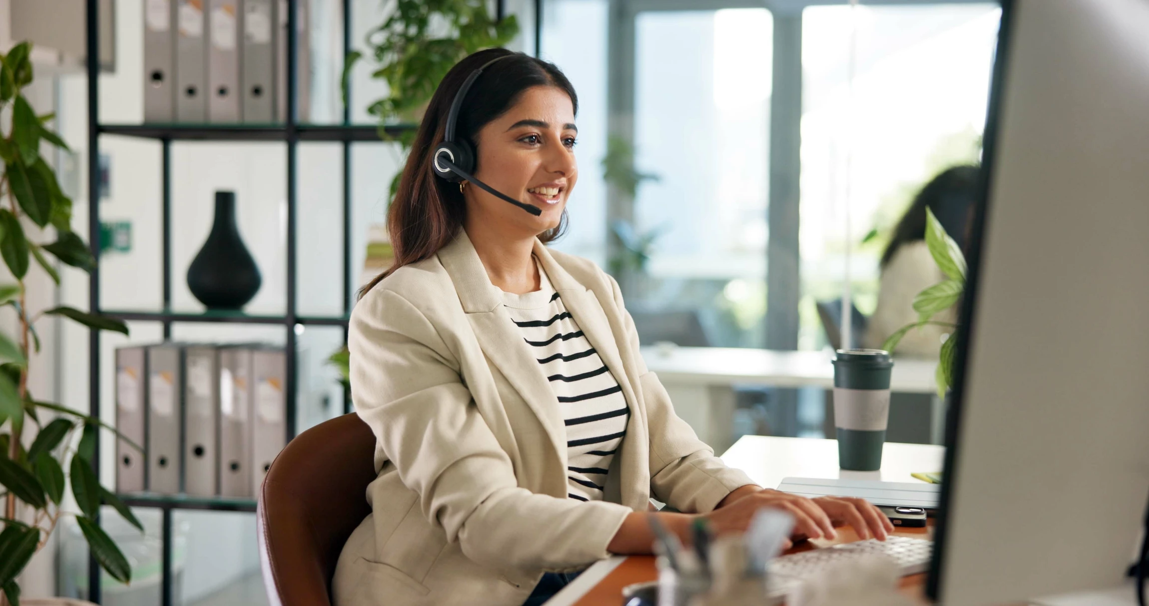 Femme souriante en chemisier blanc travaillant dans un bureau moderne, portant un casque téléphonique professionnel avec microphone-tige